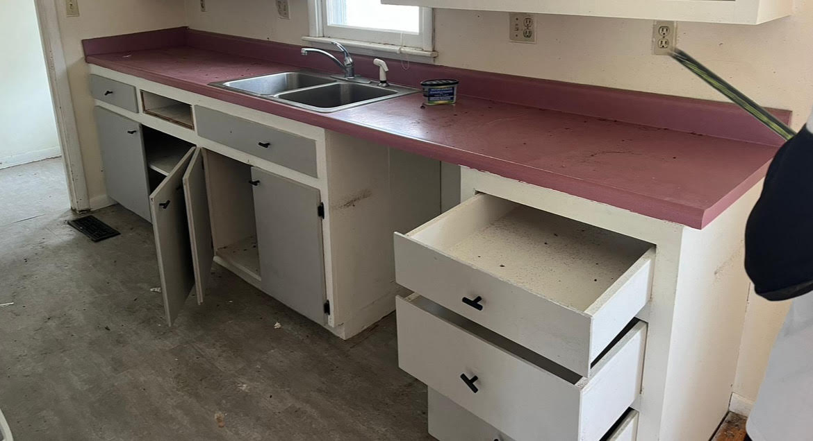 Kitchen before renovation with worn red counters, open cabinets, and unfinished flooring